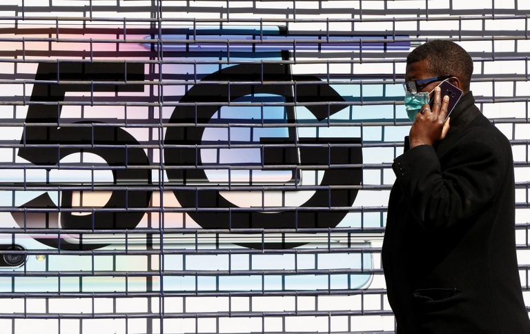 Un hombre con mascarilla hablando por su móvil, pasa delante del logo de la tecnología 5G, en una calle de Londres. REUTERS/John Sibley
